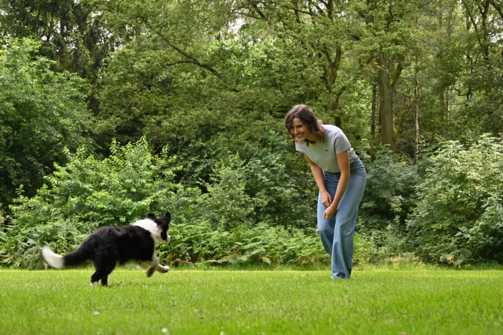 Vrouw in blauwe broek en wit shirt buigt voorover en speelt met een puppy in het gras.